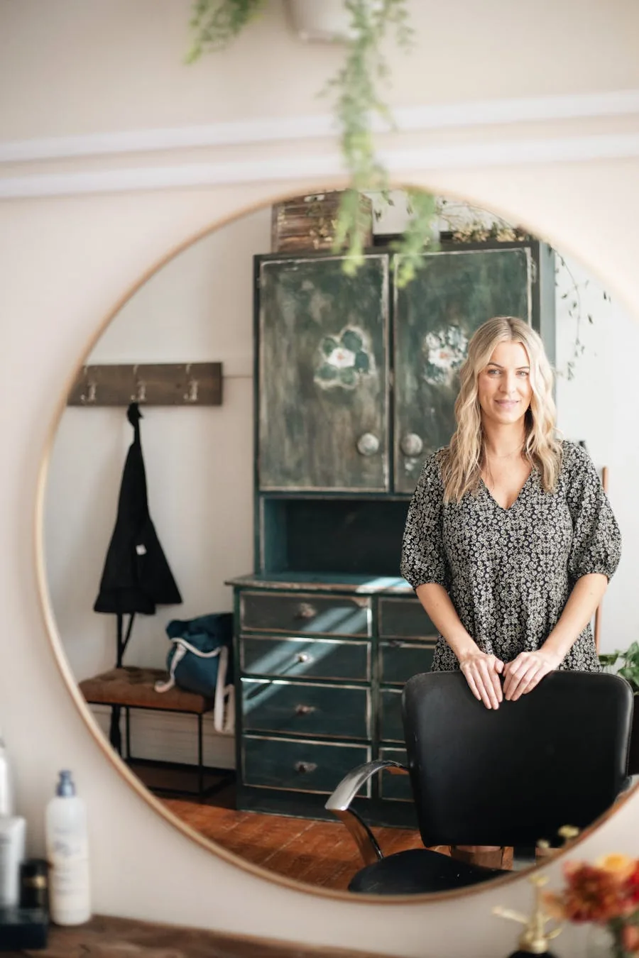 Salon professional reflected in a large round mirror inside a warm studio.